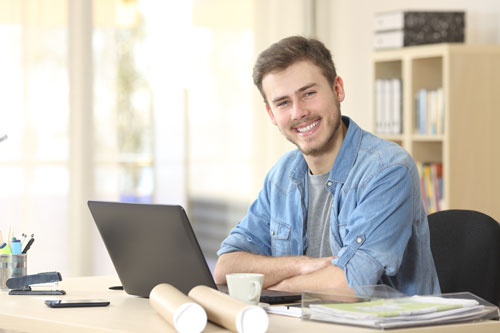 Ein junger Mann sitzt am Schreibtisch vor einem Laptop und schaut lächelnd in die Kamera