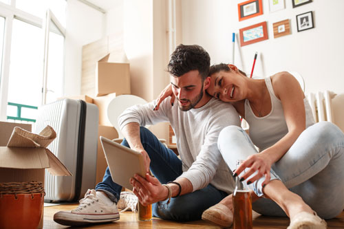 Ein glückliches junges Paar sitzt zwischen Umzugskisten auf dem Boden und nutzt einen Tablet Computer