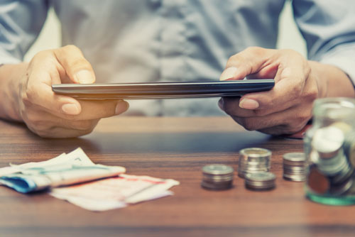Ein Mann mit Tablet Computer sitzt an einem Tisch auf dem Geldscheine und Münzen liegen